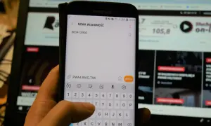 Dłoń pisze SMS na telefonie z numerem 500413900. W tle ekran komputera.