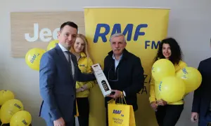 Uroczyste wręczenie nagrody w ramach finału loterii RMF FM. Mężczyzna w garniturze odbiera prezent od uśmiechniętej kobiety w żółtej kurtce.