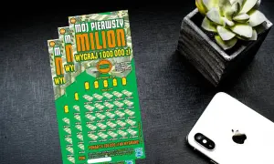 Zielona zdrapka "Mój pierwszy milion" z napisem "Wygraj 1 000 000 zł" leży obok białego iPhone'a i doniczki z sukulentem.
