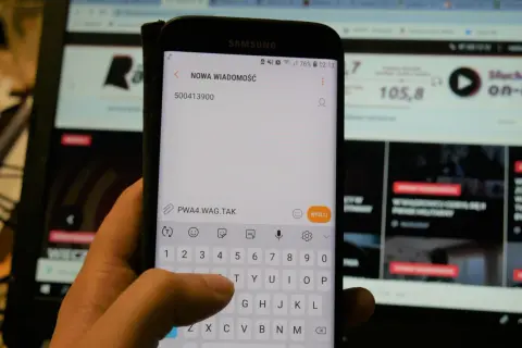 Dłoń pisze SMS na telefonie z numerem 500413900. W tle ekran komputera.