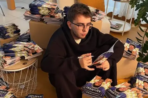Młody mężczyzna w okularach siedzi otoczony stosami czasopism z autorskimi krzyżówkami. Zarabianie na układaniu krzyżówek do czasopism to jego sposób na życie.