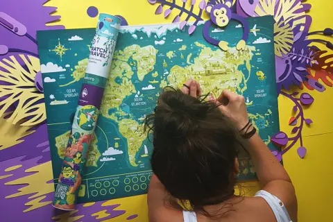 Kreatywna reklama wadliwej zdrapki bez warstwy ścieralnej: mapa świata z papierowymi dekoracjami i dziewczyną, która ją "zdrapuje".