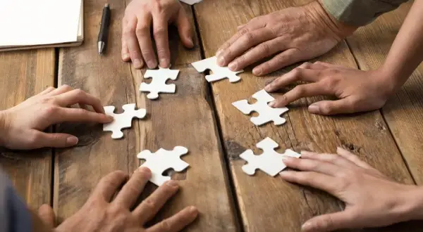 Dłonie składają puzzle, symbolizując wpływ codziennego rozwiązywania szarad na wydajność w pracy biurowej.