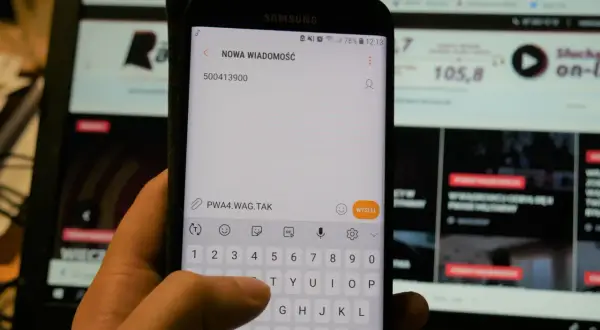 Dłoń pisze SMS na telefonie z numerem 500413900. W tle ekran komputera.