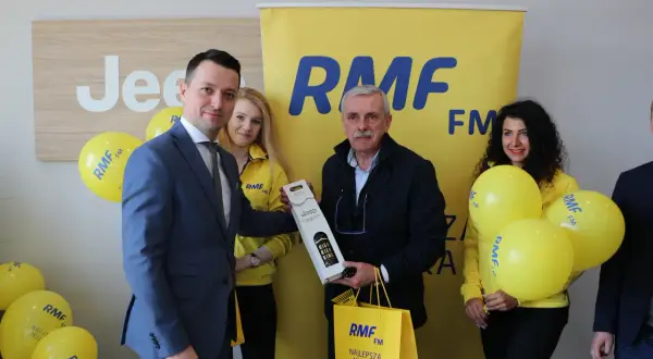Uroczyste wręczenie nagrody w ramach finału loterii RMF FM. Mężczyzna w garniturze odbiera prezent od uśmiechniętej kobiety w żółtej kurtce.