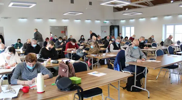 Uczestnicy Mistrzostw Polski w rozwiązywaniu krzyżówek skupieni przy stolikach, każdy z kartką i długopisem. Turniej w toku.