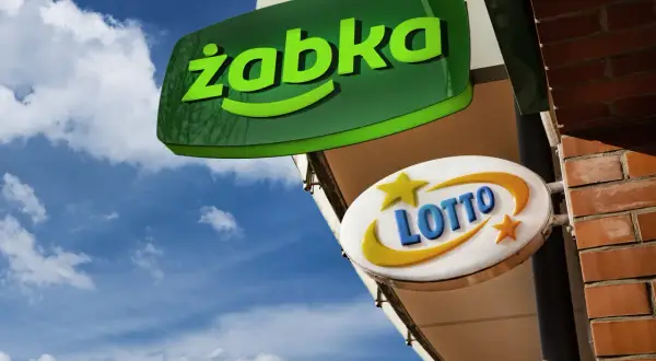Zielona tablica "Żabka" i biała z napisem "LOTTO" wiszą na tle błękitnego nieba. Czy w każdej Żabce jest lotto?