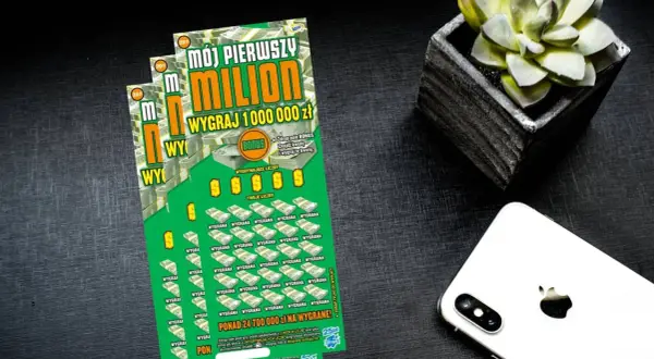 Zielona zdrapka "Mój pierwszy milion" z napisem "Wygraj 1 000 000 zł" leży obok białego iPhone'a i doniczki z sukulentem.