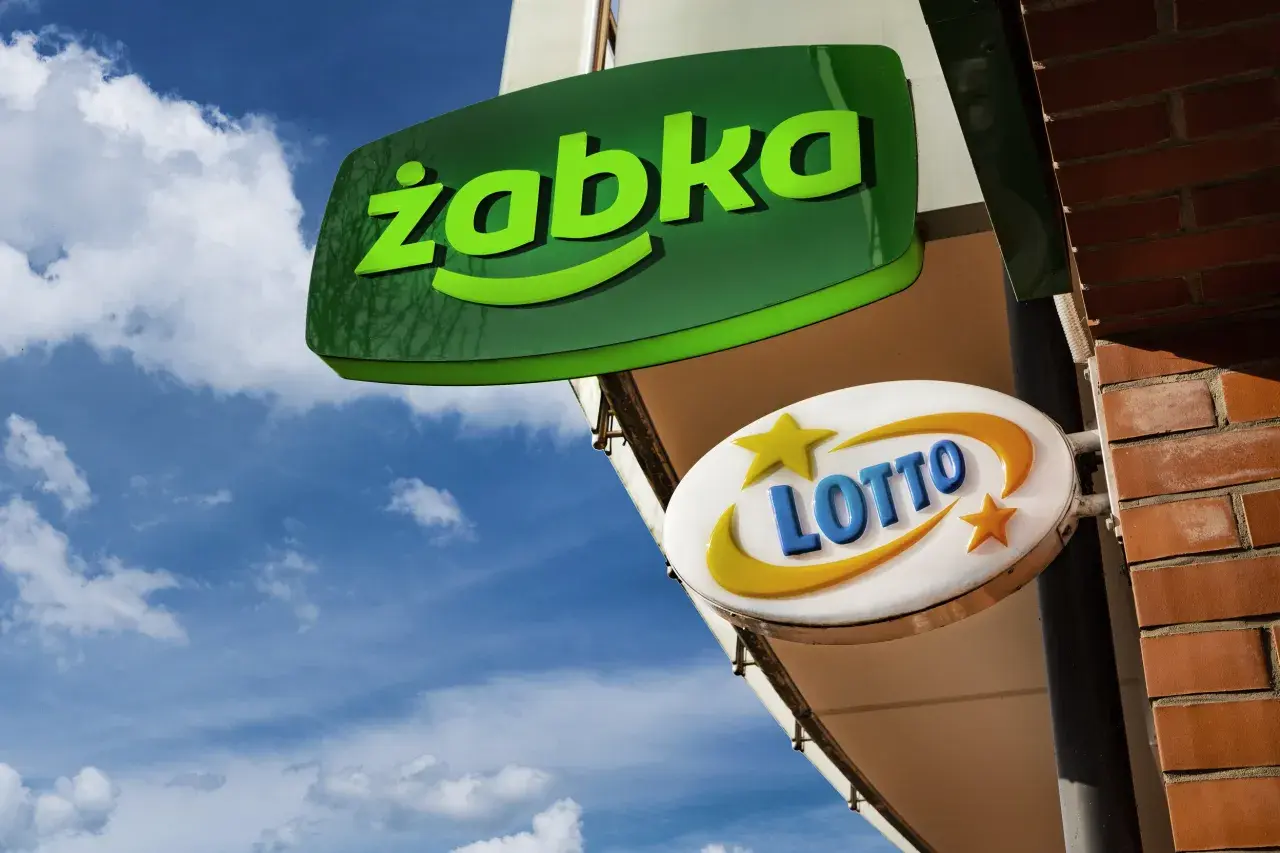 Zielona tablica "Żabka" i biała z napisem "LOTTO" wiszą na tle błękitnego nieba. Czy w każdej Żabce jest lotto?