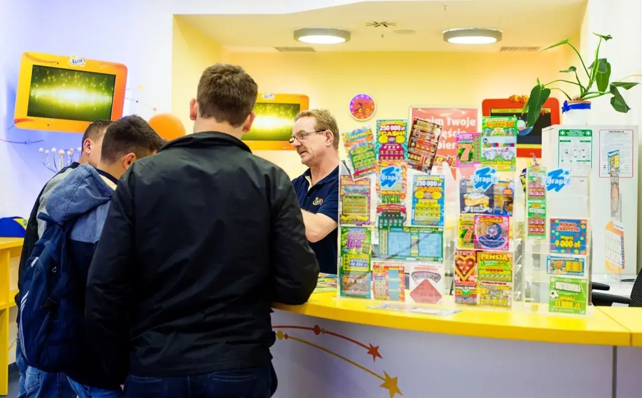 Klienci wybierają zdrapki lotto nowości przy ladzie. W tle ekran z animacją i pracownik punktu.