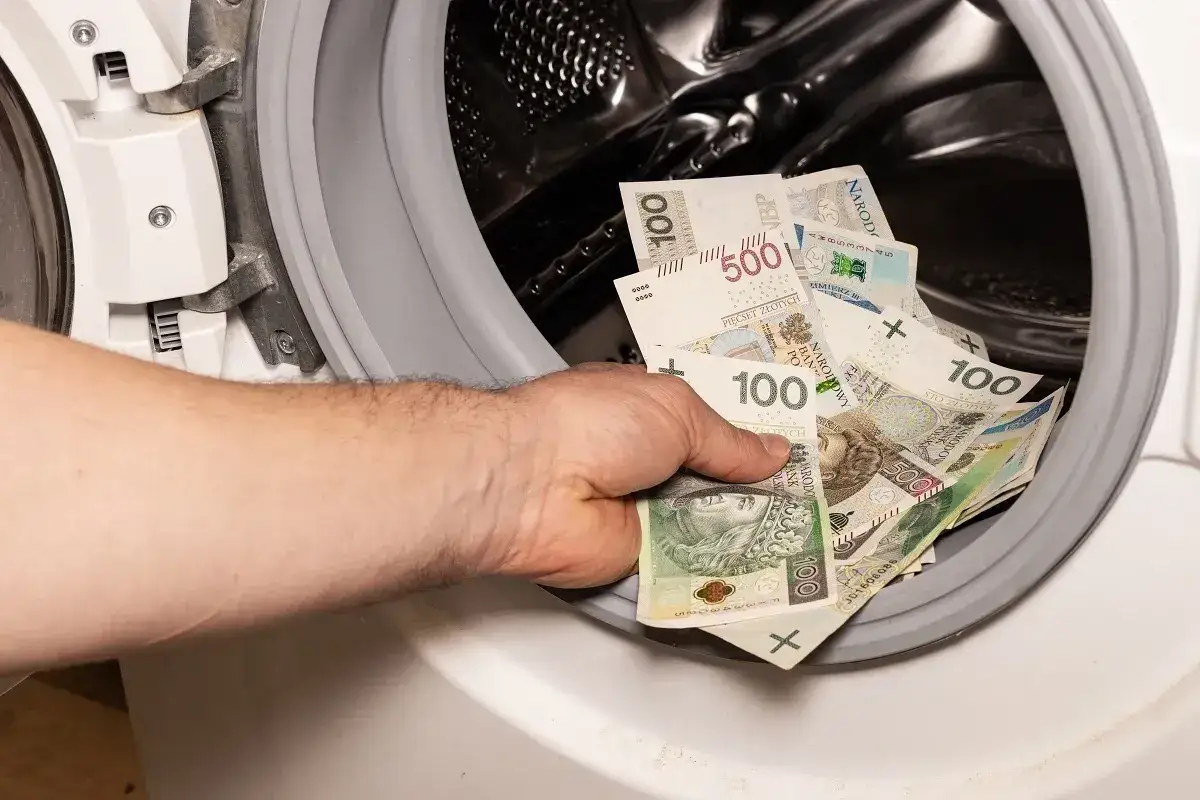 Ręka wkłada banknoty do pralki, symbolizując pranie brudnych pieniędzy przez kupony lotto, gdy system się broni.