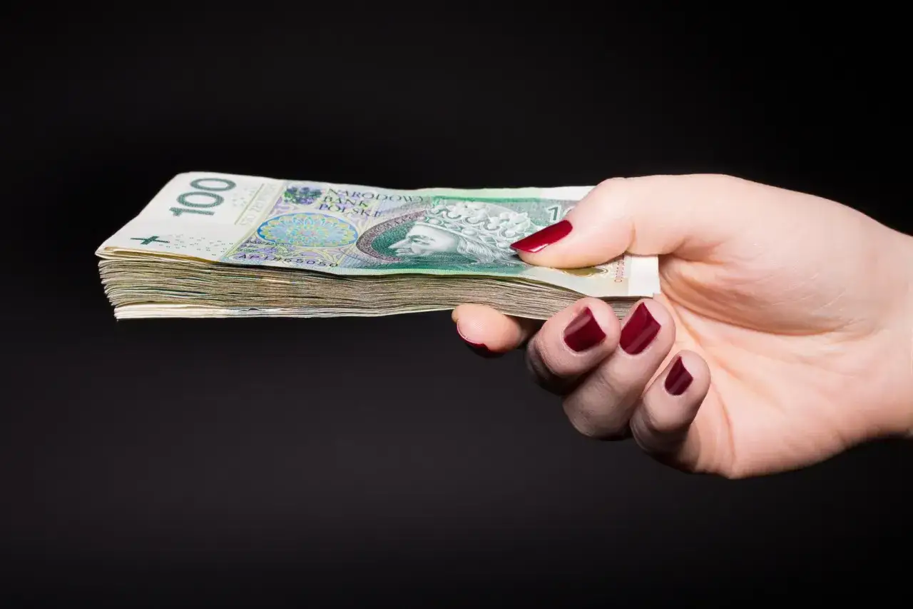 Dłoń z paznokciami pomalowanymi na czerwono trzyma plik banknotów 100 zł. Jak wyliczyć wartość dla fiskusa podatek od nagrody rzeczowej w loterii?