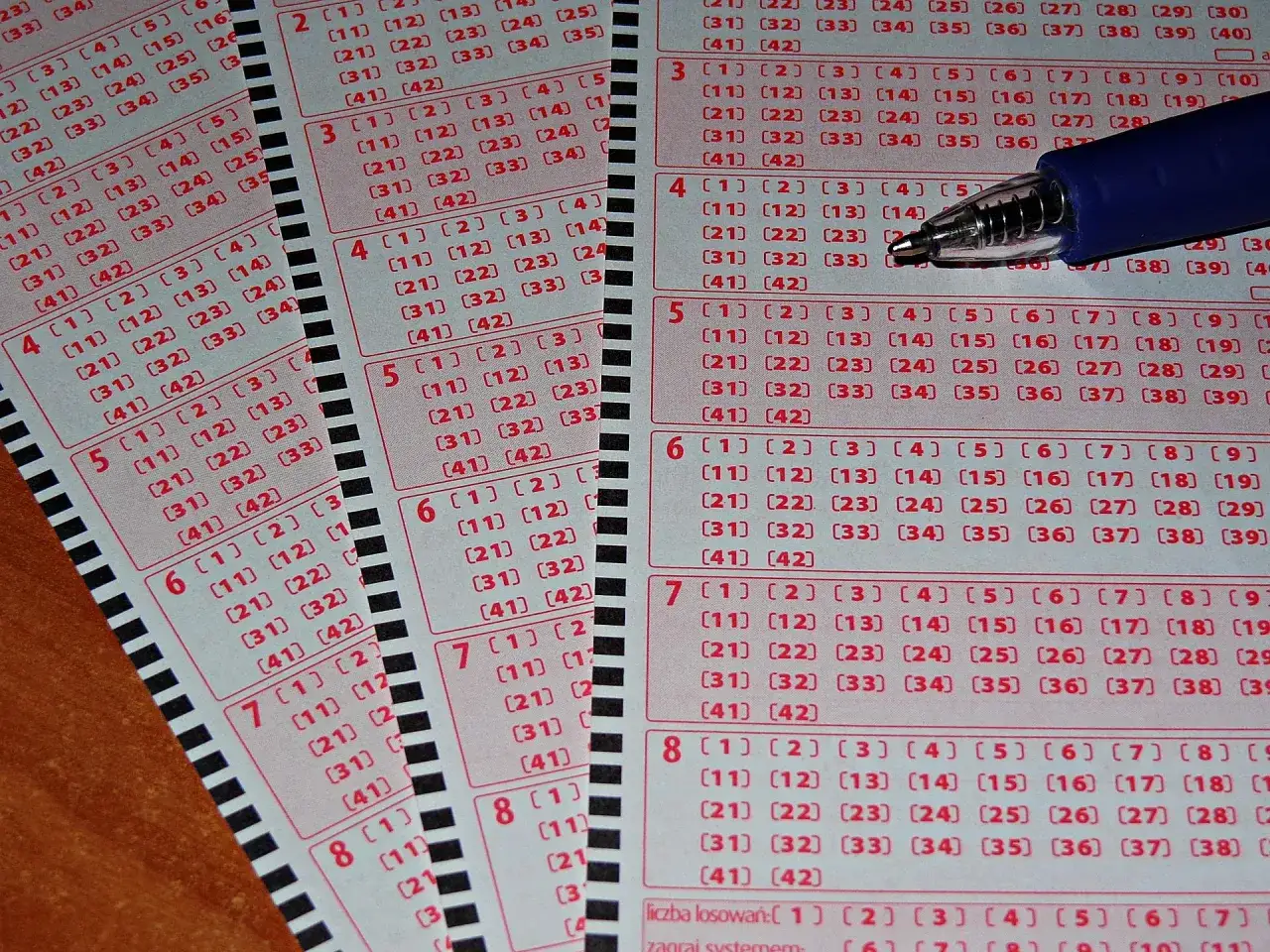 Kupony lotto z zaznaczonymi polami i długopisem. Widać dodatkowe linie i litery kontrolne, które pomagają w grze.